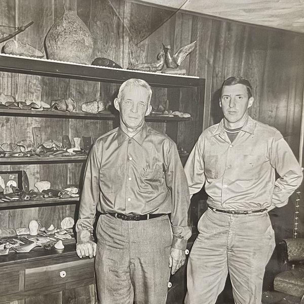 Two men stand in front of a wooden cabinet filled with pottery and artifacts.