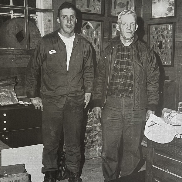 Two men standing indoors in a vintage setting with various framed pictures on the wall.